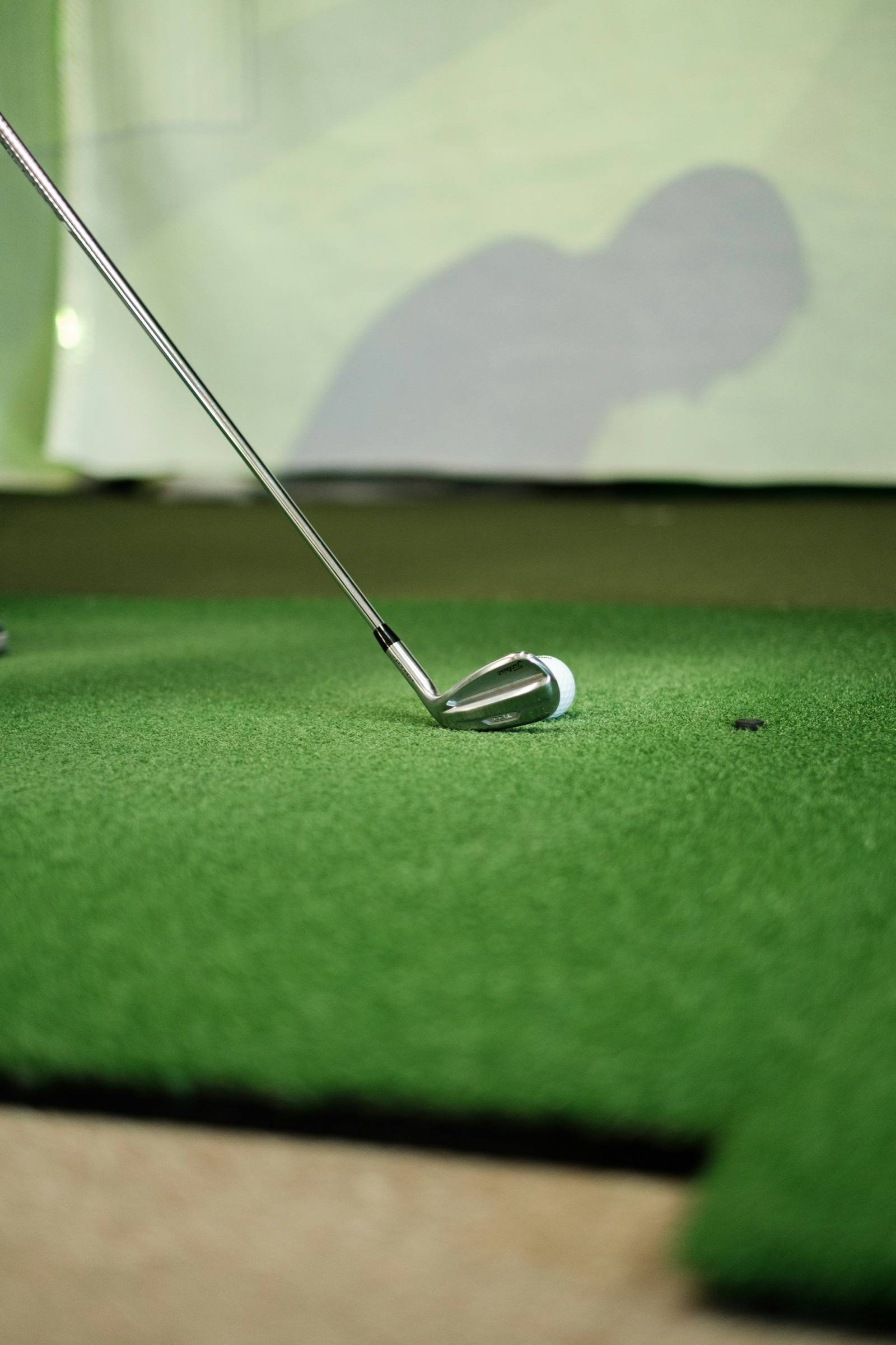 Iron addressing a ball on a hitting mat in front of an indoor golf simulator screen